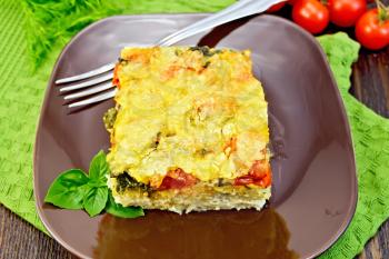 The pie of potatoes, cheese, tomato and spinach, filled egg with milk in a plate on napkin on a wooden boards background