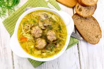Soup with meatballs, noodles and mushrooms in a white bowl on a green napkin, parsley on a background of wooden boards on top