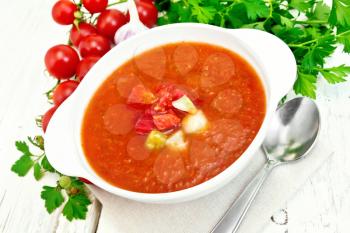Gazpacho tomato soup in a white bowl with vegetables, spoon on a napkin, garlic and parsley on a wooden board background