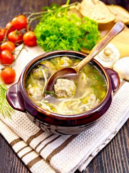Soup with meatballs, noodles and champignon in a clay bowl with spoon on a napkin, parsley, mushrooms, tomatoes and bread on a wooden boards background