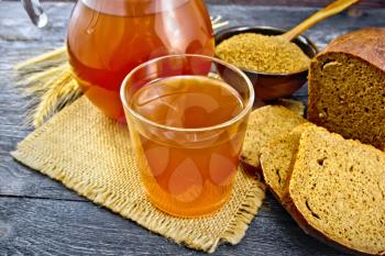 Kvass in glassful and glass jug on burlap, malt in a bowl, rye bread and ears on a wooden plank background