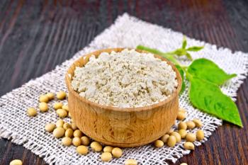 Soy flour in the bowl, soybeans on burlap, green leaf on a wooden board background