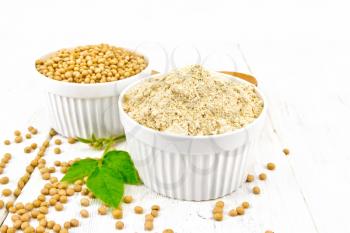 Soy flour and soybeans in two white bowls, green leaves on a wooden board background