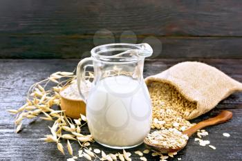 Oat milk in a jug, flour in bowl, oatmeal in a spoon, grain in bag, oaten stalks on a dark board background