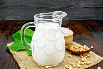 Soy milk in a jug, flour in a bowl and a fresh green leaf, soybeans in a spoon and burlap on the background of a dark wooden board