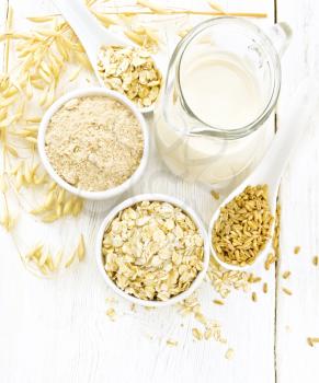 Oat flakes and flour in bowls, grain in a spoon, oatmeal milk in a glass jug and ripe oaten stalks on the background of a light wooden board from above