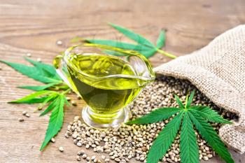 Hemp oil in a glass sauceboat with grain in a bag, leaves and stalks of cannabis against the background of an old wooden board
