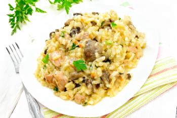 Rice risotto with mushrooms, chicken meat, cheese and garlic in a plate on kitchen towel, fork and parsley on the background of light wooden board