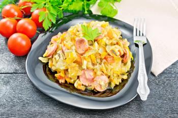 Cabbage stew with sausages in a black plate, napkin, tomatoes, parsley and fork on a dark wooden board background
