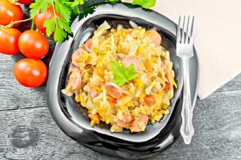 Cabbage stew with sausages in a black plate, napkin, tomatoes, parsley and fork on a wooden board background from above