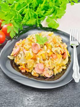 Cabbage stew with sausages in a black plate, kitchen towel, tomatoes, parsley and fork on a dark wooden board background