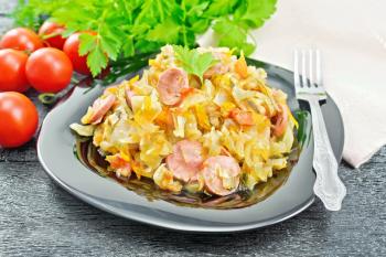Cabbage stew with sausages in a black plate, towel, tomatoes, parsley and fork on a dark wooden board background