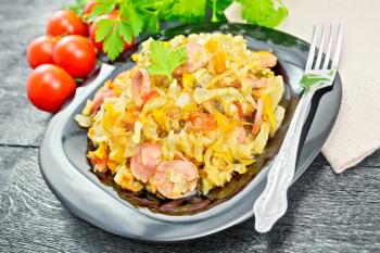 Cabbage stew with sausages in a black plate, towel, tomatoes, parsley and fork on the background of wooden boards