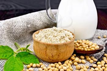 Soy flour in the bowl, soybeans in a spoon and on a napkin of burlap, milk in a jug, soya leaf on a wooden board background
