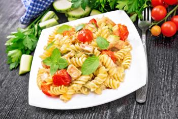 Fusilli with chicken, zucchini and tomatoes in a white plate, napkin, fork, basil and parsley on wooden board background