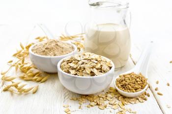 Oat flakes and flour in bowls, grain in a spoon, oatmeal milk in a glass jug and ripe oaten stalks on the background of a wooden board