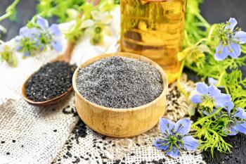 Flour of black caraway in a bowl, seeds in a spoon on burlap, oil in bottle and twigs Nigella sativa with blue flowers and leaves on wooden board background