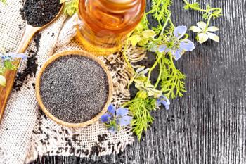 Flour of black caraway in a bowl, seeds in a spoon on burlap, oil in bottle and twigs Nigella sativa with blue flowers and green leaves on wooden board background from above