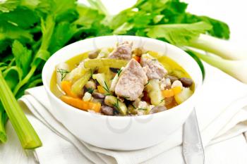 Eintopf soup of pork, celery, beans, carrots and potatoes with leek in a white bowl on a napkin on wooden board background
