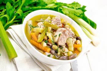 Eintopf soup of pork, celery, beans, carrots and potatoes with leek in a white bowl on a towel on wooden board background