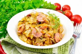 Cabbage stew with sausages in a white plate on a napkin, tomatoes, parsley and fork on a wooden board background