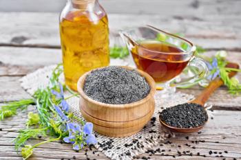 Flour of black caraway in a bowl, seeds in a spoon, oil in a bottle and sauceboat on burlap, sprigs of Nigella sativa with blue flowers and green leaves on an old wooden board