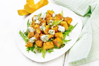 Salad of baked pumpkin, arugula with balls of salt cheese, seasoned with honey, mustard, garlic and vegetable oil in a plate, napkin and fork on white wooden board background