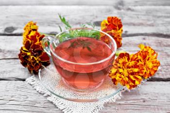 Marigold herbal tea in a glass cup and saucer on a napkin of sackcloth, fresh flowers on background of an old wooden board