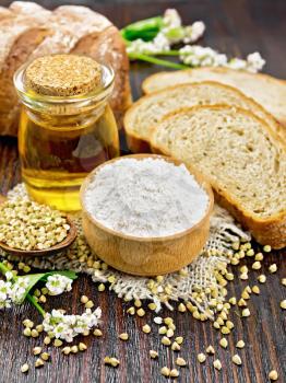 Buckwheat flour from green cereals in a bowl on sacking, buckwheat groats in a spoon and on the table, oil in a glass jar, bread, fresh flowers and leaves on dark wooden board background