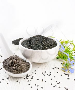Black cumin seeds in a bowl, flour and seeds in spoons, sprigs of kalingi with blue flowers and green leaves on the background of white wooden board