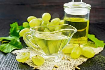 Grape oil in a gravy boat and a jar on burlap, berries of green grapes on dark wooden board background