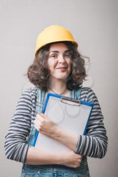 Young architect woman in yellow safety helmet holding work plan sheet, on gray background