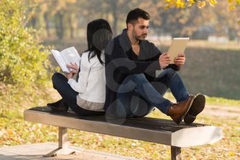 Beautiful Young Couple Sitting In The Park On A Beautiful Autumn Day - They Are Using Digital Tablet And Book