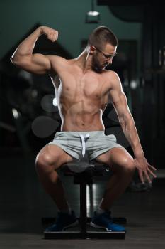 Handsome Geek Man Sitting Strong In The Gym And Flexing Muscles - Muscular Athletic Bodybuilder Fitness Model Posing After Exercises