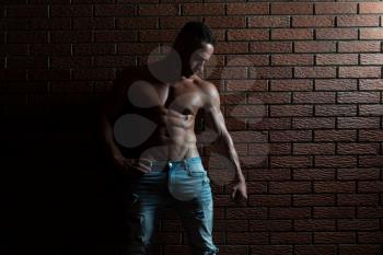 Handsome Young Man Standing Strong In The Gym And Flexing Muscles - Muscular Athletic Bodybuilder Fitness Model Posing After Exercises On Wall of Bricks