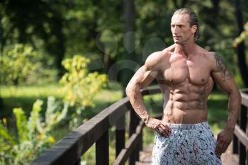 Handsome Mature Man Standing Strong Outdoors In Nature And Flexing Muscles - Muscular Athletic Bodybuilder Fitness Model Posing After Exercises