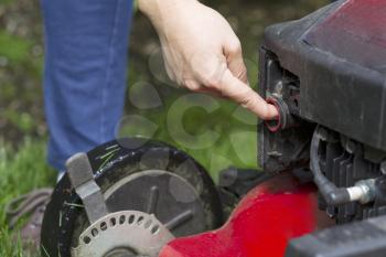 Horizontal photo of a female hand preparing to start an old gas lawnmower on grass yard with partial blue jeans, shoe and green grass in background