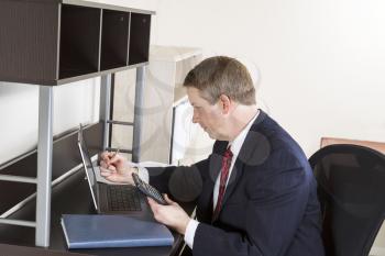 Mature man holding calculator and pen while checking numbers on computer with papers on desk