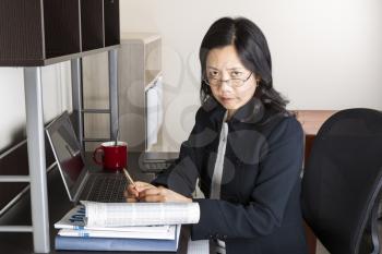 Professional Mature Asian woman with glasses down while working on income taxes with tax form booklet, calculator, coffee cup and computer on desk
