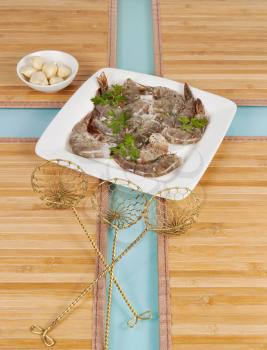 Vertical layout of fresh large raw shrimp with skin on, parsley, peeled garlic and food strainers on natural bamboo place mats with green glass table in background