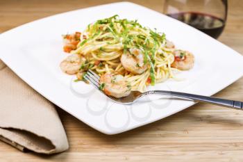 Closeup horizontal photo of Pasta dish with focus on large shrimp in stainless steel fork, basil, cloth napkin, red wine, and parsley inside of white dish