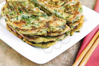 Horizontal photo of stacked Korean green onion pancakes in plate, chopsticks on cloth napkin with stone table underneath