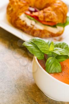 Vertical photo, slightly angled, of fresh basil in tomato soup in bowl with chicken sandwich in background all resting on natural stone counter top 