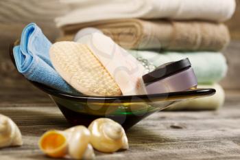 Horizontal eye level view of spa hygiene accessories in glass bowl with clean stacked towels in background and seashells on rustic wood