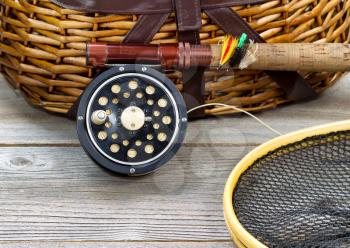 antique fly fishing reel, rod, landing net, creel and artificial flies on rustic wood. Layout in horizontal format.