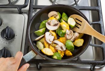 Close up of hand holding frying pan, focus on food, while stirring vegetables with wooden spoon.