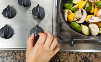 Close up of hand turning on stove with vegetables in frying pan on side. 