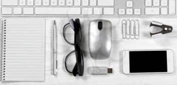 Overhead view of a white desktop with basic office stationery objects lined up under keyboard.   