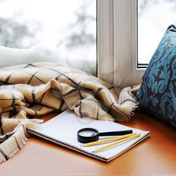 Open notepad, magnifier glass, pillow, pencils and beige warm plaid located on stylized wooden windowsill. Winter concept of comfort and relaxation.