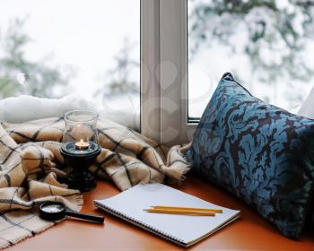 Open notepad, magnifier glass, pillow, candle, pencils and beige warm plaid located on stylized wooden windowsill. Winter concept of comfort and relaxation.